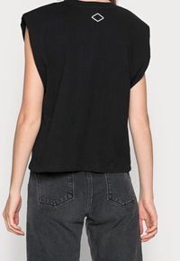 Osoba w czarnej, bezrękawnikowej bluzce z małym białym logo w kształcie diamentu na górnej części pleców oraz w wyblakłych czarnych dżinsach, pokazana z tyłu.