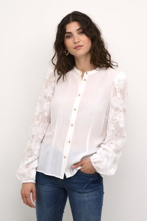 Camicia bianca con maniche lunghe trasparenti ricamate, chiusura con bottoni, scollatura rotonda e spalle leggermente gonfie. Abbinata a jeans blu.