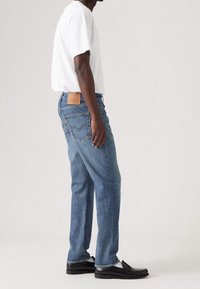 Jeans de denim azul con un diseño de pierna recta, usados con una camiseta blanca, que cuentan con un parche de cuero marrón en la parte trasera de la cintura y zapatos negros.