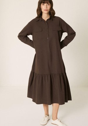 Γυναίκα που φοράει μακρυμάνικο, σκούρο καφέ, midi φόρεμα με κουμπιά και τσέπες, συνδυασμένο με λευκά αθλητικά παπούτσια, στέκεται με τα χέρια στη μέση.