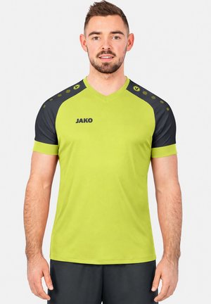 Mann trägt limegrünes und schwarzes Sportshirt mit kurzem Arm und „JAKO“-Logo, steht vor einem einfarbigen hellen Hintergrund.