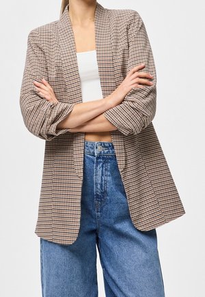 Femme portant un blazer marron à grands carreaux pied-de-poule sur un crop top blanc et un jean bleu taille haute, les bras croisés devant elle.
