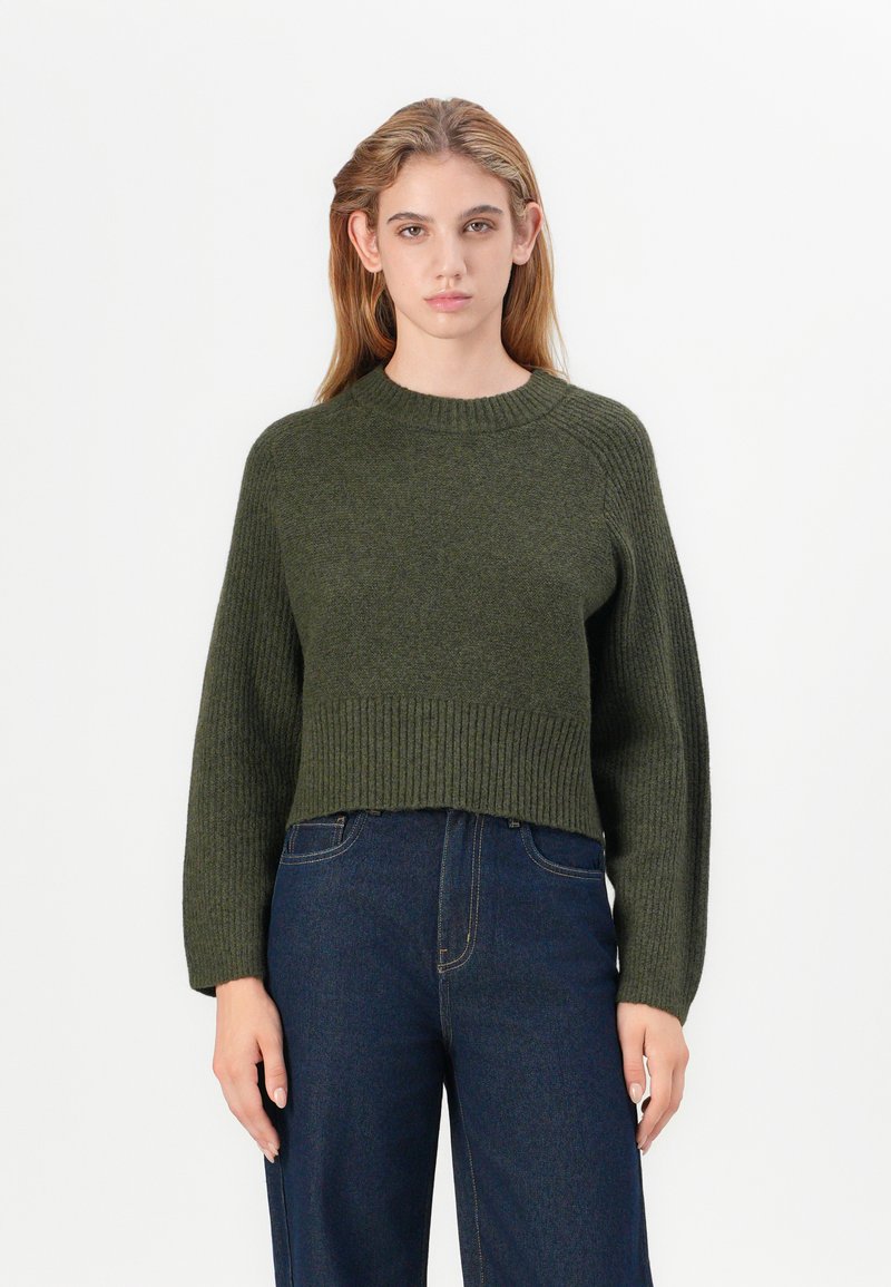 Maglione verde corto con scollo e orlo a costine, maniche lunghe e motivo a maglia testurizzato, abbinato a jeans scuri a vita alta.