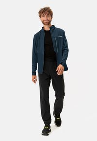 Donkerblauwe heren performance jack met een gladde textuur, lange mouwen, opstaande kraag en een ritszak, gecombineerd met zwarte broek en sneakers.