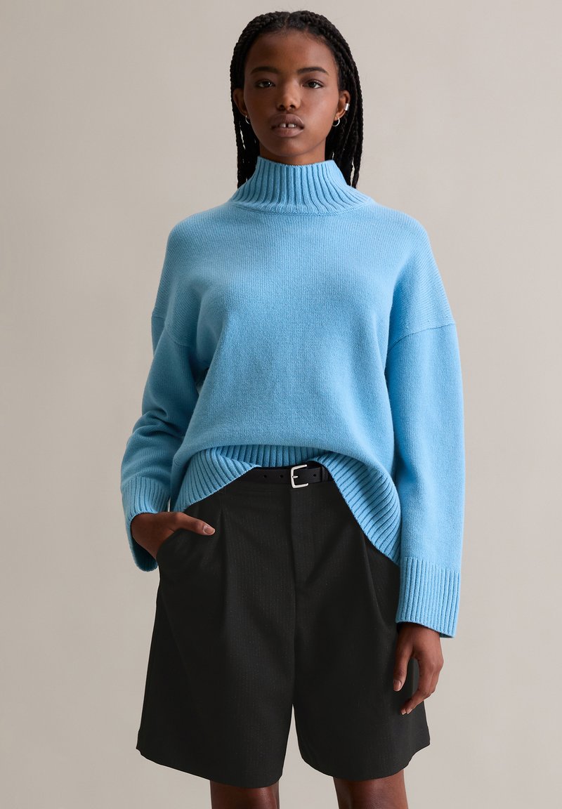 Lichtblauwe gebreide trui met een hoge geribde kraag en verwijde schouders, gecombineerd met zwarte plooishorts en een zwarte riem.