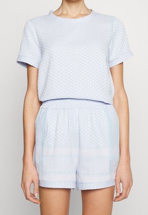 Femme portant un ensemble assorti composé d'un haut à manches courtes texturé bleu clair et d'un short avec taille élastique, debout devant un fond uni.