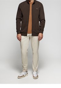 Hnědá zipová bunda přes béžové tričko, doplněná světle barevnými džíny a bílými teniskami s hnědými akcenty. Jednoduchý a ležérní design.