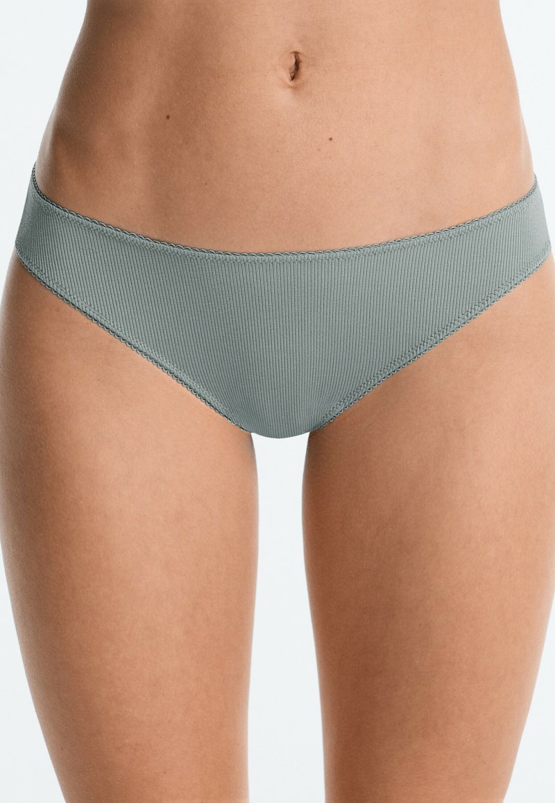 Femme portant un sous-vêtement bikini côtelé gris clair, montrant le ventre et le haut des cuisses sur un fond clair uni.