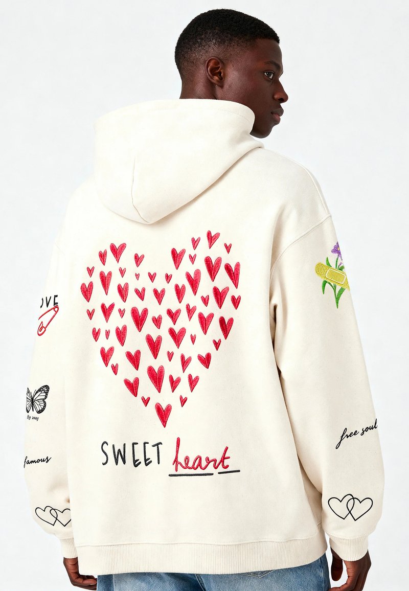 Hombre que lleva una sudadera color crema con corazones rojos formando un gran corazón y el texto "Dulce corazón", además de flores y corazones bordados en las mangas.