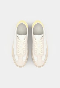 White sneakers with beige suede accents and a yellow rubber heel cap. Features flat laces, a smooth texture, and a rounded toe shape.