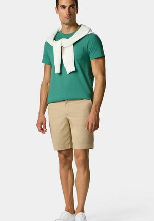 Zelené bavlněné tričko s kapsou, béžové kraťasy a bílé svetr zavázaný kolem krku; bílí tenisky dotvářejí celý outfit.