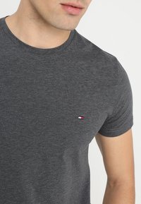 T-shirt grigio antracite a girocollo in cotone, con un piccolo logo rosso, bianco e blu sul petto sinistro. Maniche corte, superficie liscia.