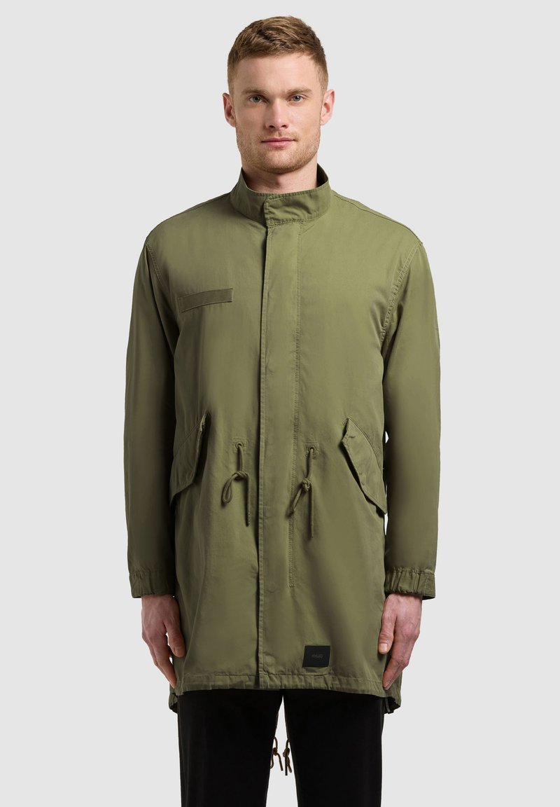 Homme portant une longue veste vert olive avec poches avant et taille ajustable par cordon, debout devant un fond clair uni.