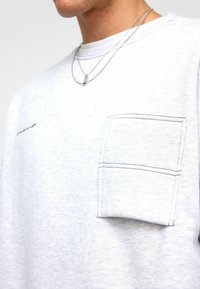 Sweat-shirt gris clair en mélange de coton. Présente une poche sur la poitrine gauche avec des coutures contrastées et un col rond. Deux chaînes argentées superposées.