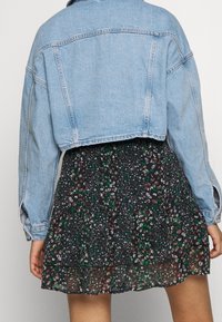 Bakåtsikt av en person som bär en ljusblå cropped denimjacka över en svart blommönstrad kjol med gröna och bruna detaljer.