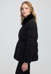 Zwarte pufferjack met een nep bontkraag, met een aansluitende taille, zijzakken en een geborduurde textuur. Effen achtergrond, model kijkt naar rechts.