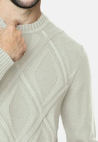 Suéter de punto de color beige claro con un cuello acanalado y patrones de diamante texturizados, que presenta mangas largas y una textura suave y gruesa.