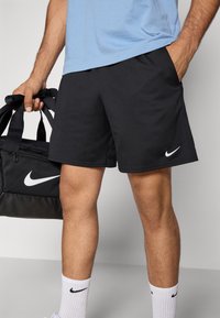 Μαύρο σόρτς Nike με ελαστική μέση και πλαϊνές τσέπες. Συνδυασμένο με ανοιχτό μπλε πουκάμισο και ντεκόρ από μαύρη τσάντα duffel Nike.