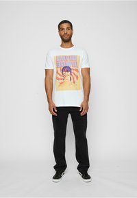 Hvid bomulds t-shirt med et farverigt David Bowie grafik. Modellen parrer den med sorte jeans og sorte sneakers med hvide såler.