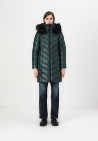 Cappotto verde puffoso con colletto alto e cappuccio in pelliccia sintetica nera, caratterizzato da un motivo a rombi e una zip frontale. Indossato con jeans scuri.