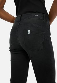 Jean en denim noir au design ajusté, présentant un logo sur la taille et un petit accent métallique sur la poche arrière.