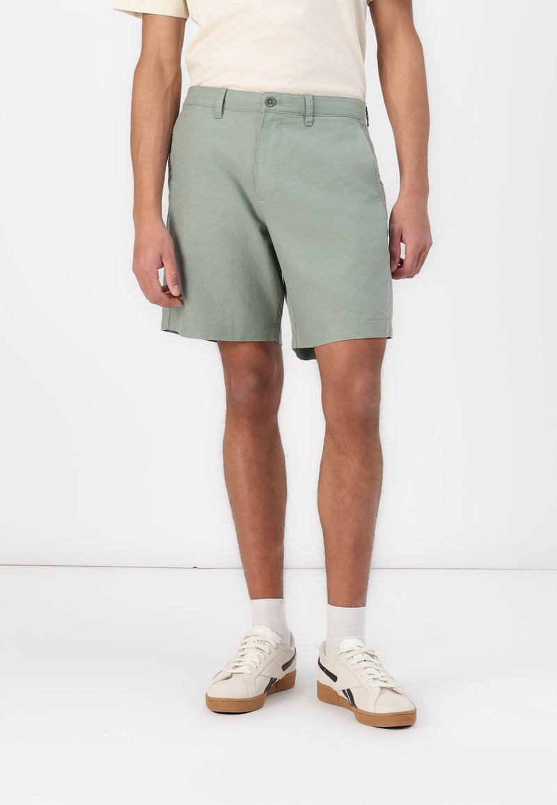 Shorts en coton verts, de longueur mi-cuisse, avec une fermeture à bouton et des passants de ceinture. Portés avec des baskets blanches et des chaussettes de longueur crew. Design simple.
