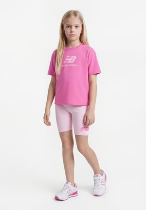 Ragazza giovane in piedi che indossa una maglietta rosa New Balance, pantaloncini da ciclismo rosa chiaro e scarpe da ginnastica bianche con dettagli rosa.