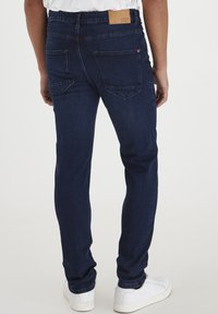 Jeans de mezclilla azul oscuro con un corte ajustado, que cuentan con bolsillos traseros y un parche de cuero en la cintura, combinados con zapatillas blancas.