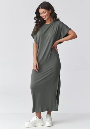 Kobieta z długimi kręconymi włosami w luźnej, szarej, maxi sukience i białych trampkach, uśmiechająca się z jedną ręką na biodrze.