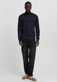 Maglione a collo alto blu navy in una trama a maglia, abbinato a jeans scuri e sneakers marroni con strisce bianche, in piedi su uno sfondo neutro.