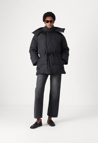 Black puffer jacket with a cinched waist, high collar, and large pockets, paired with dark, wide-leg jeans and black loafers.