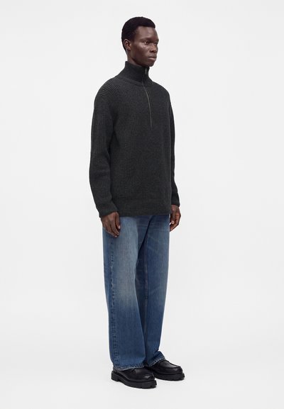 Homme portant un pull gris foncé à fermeture éclair partielle, un jean bleu ample et des chaussures noires, debout devant un fond blanc uni.