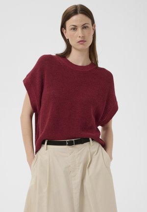 Femme aux cheveux bruns raides portant un haut en tricot bordeaux sans manches, rentré dans un pantalon beige taille haute à plis avec une ceinture noire.