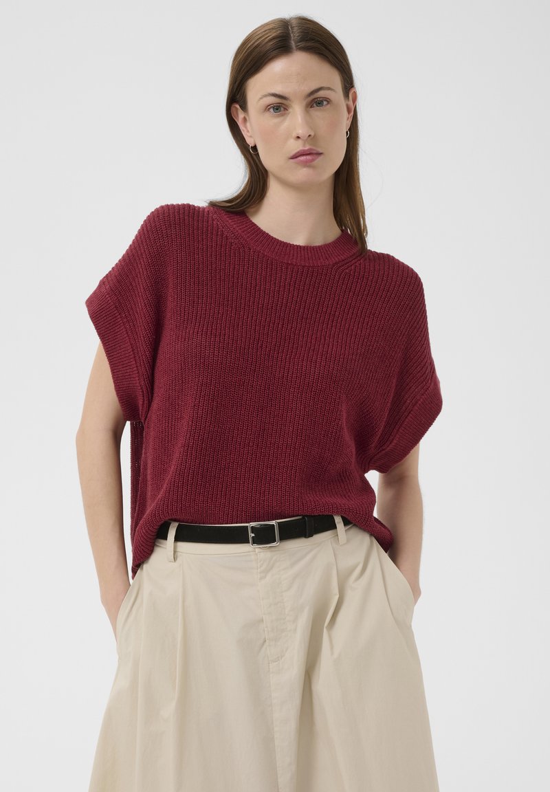 Femme aux cheveux bruns raides portant un haut en tricot bordeaux sans manches, rentré dans un pantalon beige taille haute à plis avec une ceinture noire.