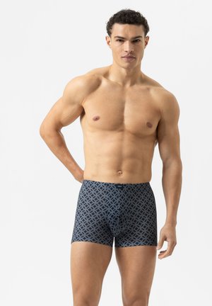 Spierachtige man in donkere boxerondergoed met een in elkaar grijpende grijze cirkelpatroon, staand tegen een effen witte achtergrond.