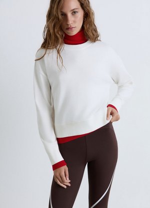 Donna con capelli ricci che indossa un maglione bianco sopra un dolcevita rosso e leggings marroni con strisce curve bianche su uno sfondo uniforme.