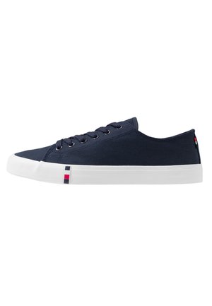 Zapatilla de lona azul marino de perfil bajo con suela de goma blanca y pequeños detalles cuadrados rojos y azul marino cerca de la puntera y el talón.