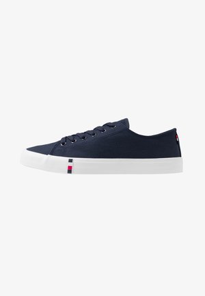 Zapatilla de lona azul marino de perfil bajo con suela de goma blanca y pequeños detalles cuadrados rojos y azul marino cerca de la puntera y el talón.