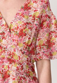 Květinové šaty s růžovým pozadím, na kterém jsou červené a zelené květy, V-výstřihem, krátkými rukávy a řaseným detailem v pase.