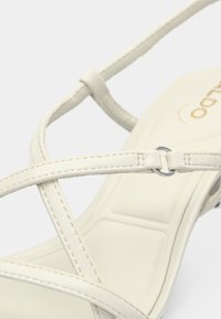 Sandale beige à lanières croisées avec un accent circulaire argenté. Matière en simili cuir lisse avec une semelle intérieure rembourrée. Design classique.