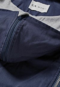 Námořnicky modrá bunda na zip s šedou podšívkou kapuce. Má hladkou tkaninu a kontrastní zip. Uvnitř je viditelná značka "CA MAMA".