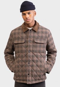 Mand iført brun quilted plaid jakke med corduroy krave og sort hue, stående med hænderne i lommerne mod en hvid baggrund.