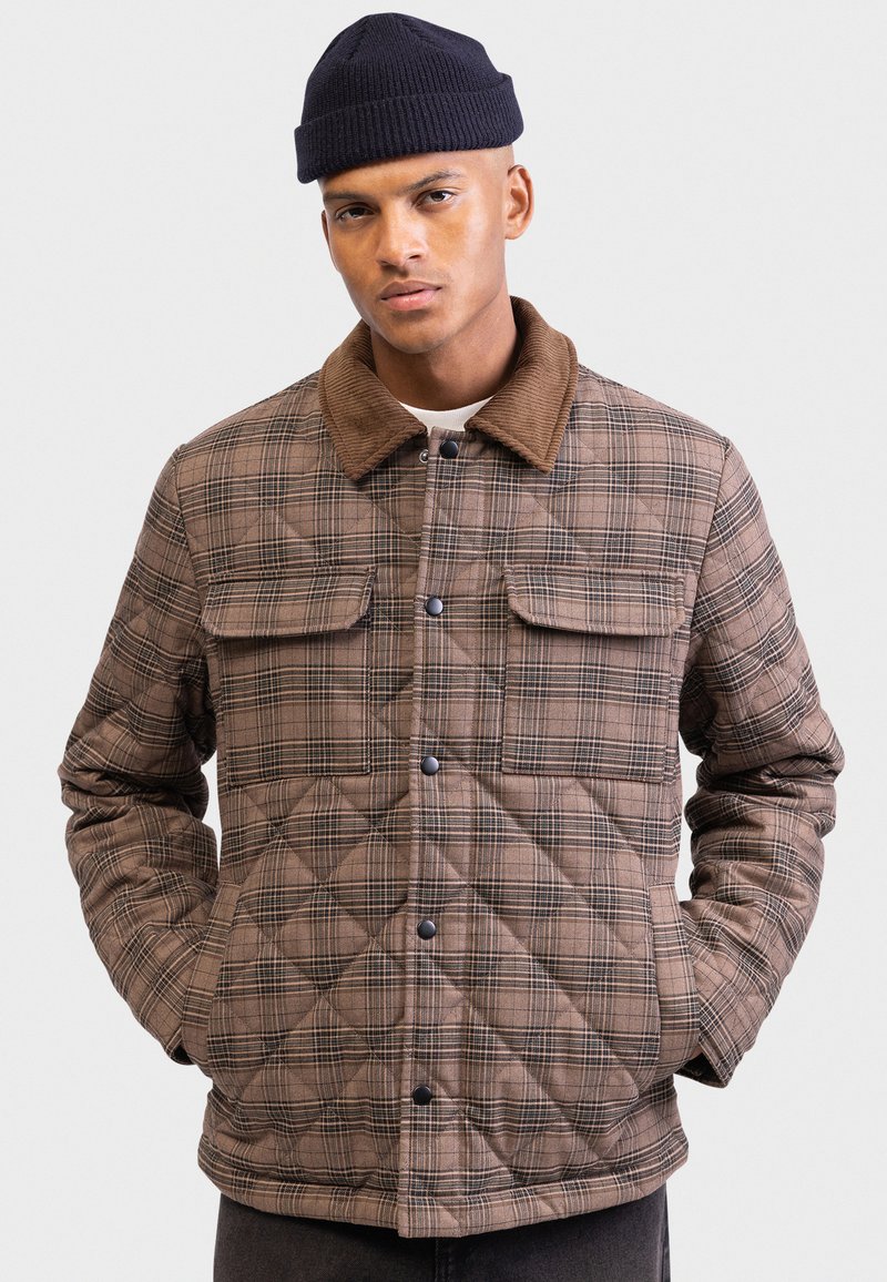 Mand iført brun quilted plaid jakke med corduroy krave og sort hue, stående med hænderne i lommerne mod en hvid baggrund.