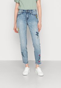 Jeans de mezclilla azul claro con flores bordadas en amarillo, rosa y azul, con dobladillos deshilachados, combinados con zapatillas blancas y una camisa verde.