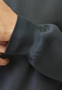 Tamno siva man�žeta dukserice s rebrastom teksturom, koja prikazuje mekanu tkaninu i uredne šavove. Prikaz u blizini dizajna pripijenog rukava.