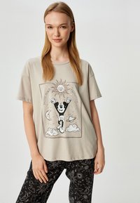 Oversized beige katoenen t-shirt met een grafische print van een personage met een zon en hemelse elementen, binnen een omrand ontwerp.