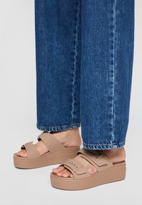 Ljusbruna plattformssandaler med breda remmar; tillverkade av mjukt material; har små nitar och en tjock sula. Bärs med blå denim.