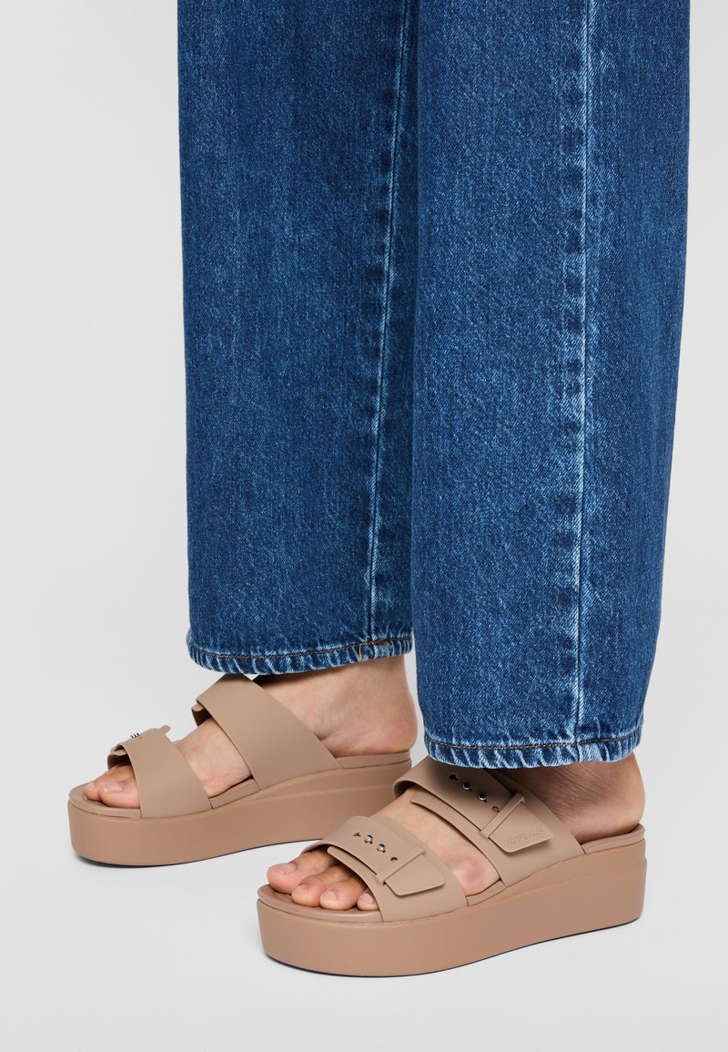 Ljusbruna plattformssandaler med breda remmar; tillverkade av mjukt material; har små nitar och en tjock sula. Bärs med blå denim.