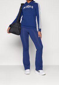 Osoba v modrém adidas teplákovém kostýmu s bílými pruhy, bílých adidas teniskách a nese černou sportovní tašku přes jedno rameno.