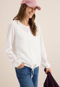 Blusa blanca de manga larga con dobladillo ajustable, hecha de tela ligera. Combinada con jeans de mezclilla azules y una gorra de pana rosa.